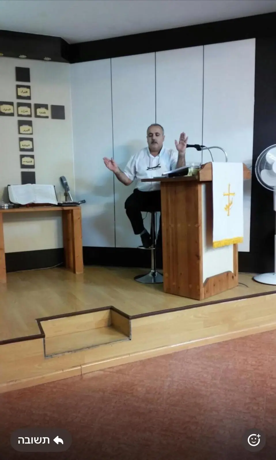 A teacher seated on a stool on the platform, teaching the congregation with open hands